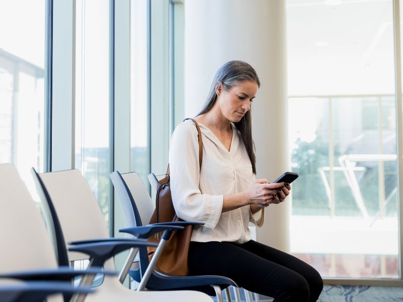 Een vrouw zit op een rij stoelen. Ze draagt een witte blouse en een bruine tas over haar schouder, en kijkt geconcentreerd naar de digitale Persoonlijke Gezondheidsomgeving op haar telefoon.